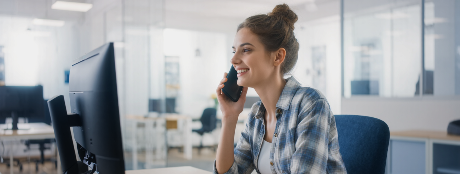 junge Frau telefoniert mit Handy an Schreibtisch vor Monitor